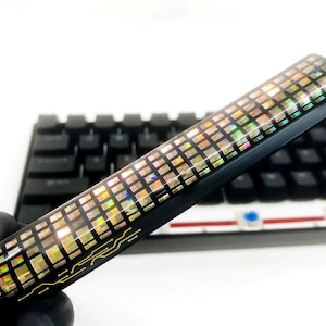 Può includere: Un oggetto rettangolare nero e iridescente con un motivo a griglia. L'oggetto ha un design dorato su un'estremità e viene tenuto davanti a una tastiera nera. La griglia riflette più colori.