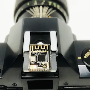 May include: Close-up of a black camera with a small circuit board attached to the top. The circuit board has gold accents and the text "LYBR1S3 V1.0". The camera has a dial labeled "SELF PROGRAM".