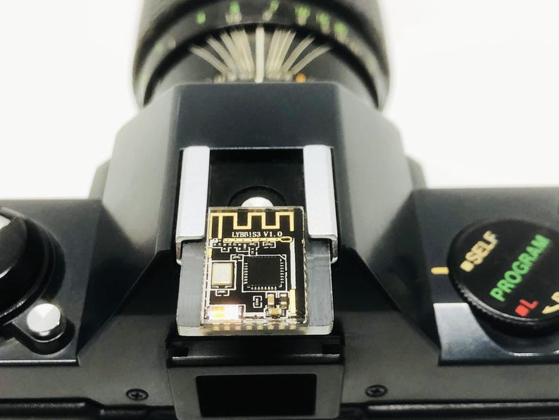 May include: Close-up of a black camera with a small circuit board attached to the top. The circuit board has gold accents and the text "LYBR1S3 V1.0". The camera has a dial labeled "SELF PROGRAM".