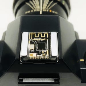 May include: Close-up of a black camera with a circuit board insert. The circuit board has gold and black details, with the text "LYBR153 V1.0". The camera has a dial with "SELF" and "PROOF" markings.