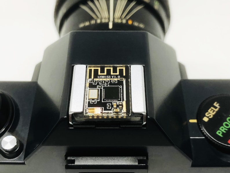 May include: Close-up of a black camera with a circuit board insert. The circuit board has gold and black details, with the text "LYBR153 V1.0". The camera has a dial with "SELF" and "PROOF" markings.