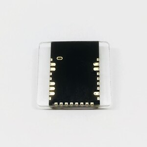May include: A small, rectangular electronic component with a black center and gold-colored pins. The component is encased in a clear, square plastic housing. The number "0" is visible on the black surface.