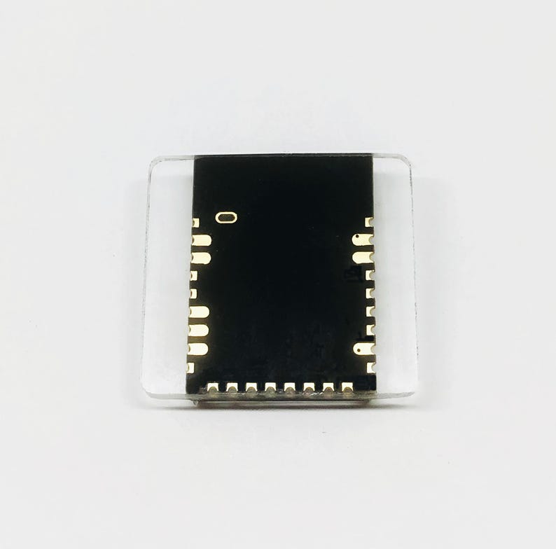 May include: A small, rectangular electronic component with a black center and gold-colored pins. The component is encased in a clear, square plastic housing. The number "0" is visible on the black surface.