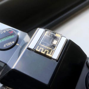 May include: Close-up of a black camera with a silver top and a small circuit board. The circuit board has a clear plastic cover and gold-colored accents. The camera has a dial with the word "SELF" on it.