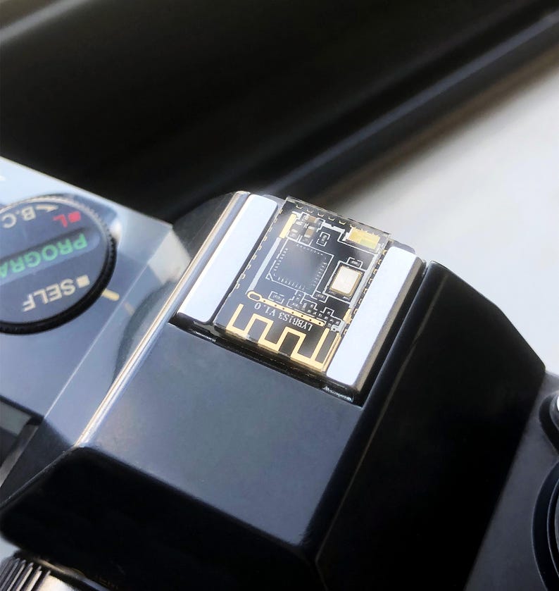 May include: Close-up of a black camera with a silver top and a small circuit board. The circuit board has a clear plastic cover and gold-colored accents. The camera has a dial with the word "SELF" on it.