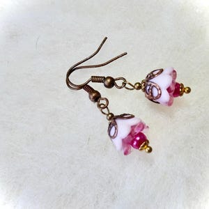 May include: A pair of dangle earrings featuring white and pink flower-shaped beads. The earrings have bronze-colored hooks and accents, with small gold-colored beads. The floral beads have a translucent quality.