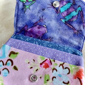 May include: A small, open fabric wallet with a snap closure. The interior features layers of patterned fabric in shades of purple, blue, and pink. The top flap is a blue fabric with frog illustrations in purple and teal.