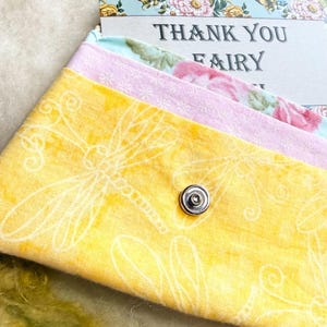 May include: A fabric envelope-style pouch featuring a yellow dragonfly print, a light pink middle section, and a floral patterned flap with a metal snap closure. A card inside reads THANK YOU FAIRY.