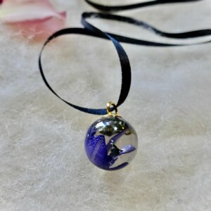 May include: A clear orb pendant with a gold bail, filled with blue floral elements. The pendant hangs from a black ribbon necklace. The background is a soft, white, textured surface.