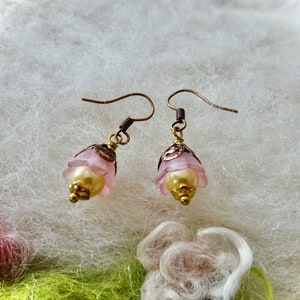 May include: A pair of delicate earrings featuring a light pink flower design with a gold-colored bead center. The earrings have a vintage aesthetic with bronze-colored hooks and accents, set against a soft, textured background.