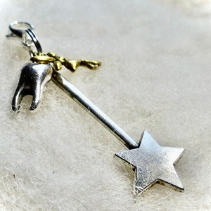 May include: A silver-colored charm featuring a tooth, a star, and a small gold-colored key. The charm is attached to a clasp, and is likely a pendant or a charm for a bracelet.