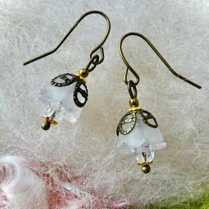 May include: A pair of delicate earrings featuring frosted white bell-shaped flowers with intricate bronze filigree caps and clear crystal accents. The earrings have bronze-colored ear wires and small gold-colored beads.