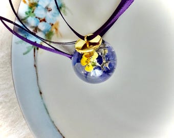 May include: A gold pendant necklace with a clear glass orb containing pressed purple and yellow flowers. The necklace is on a purple ribbon.
