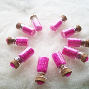 May include: Ten small glass bottles filled with pink glitter, arranged in a circle on a white fluffy surface.