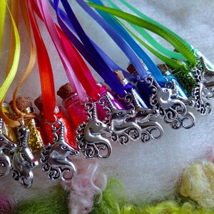 May include: A selection of miniature glass bottles filled with coloured glitter, each with a cork stopper. Silver unicorn charms hang from rainbow-coloured ribbons. The bottles are presented on a soft, white surface.