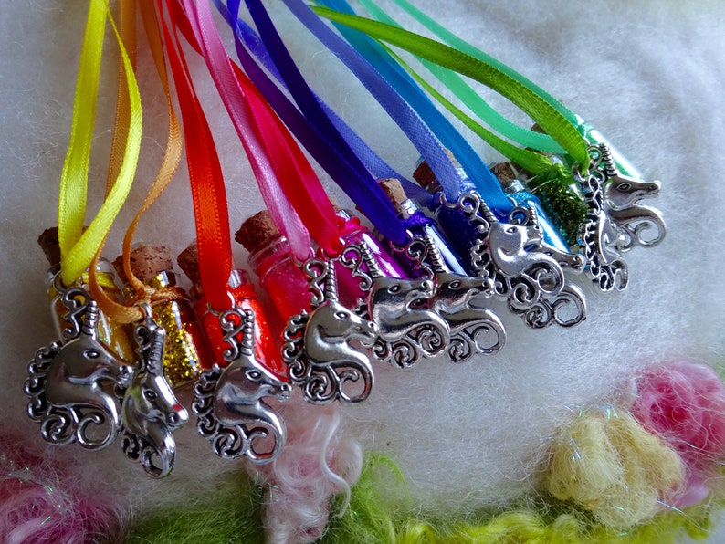May include: A selection of miniature glass bottles filled with coloured glitter, each with a cork stopper. Silver unicorn charms hang from rainbow-coloured ribbons. The bottles are presented on a soft, white surface.