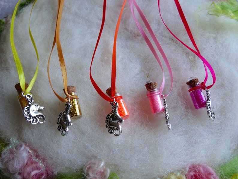 May include: A selection of miniature glass bottles filled with coloured glitter, each suspended by a ribbon. The bottles are decorated with silver unicorn charms. Ribbons are yellow, orange, red, pink, and fuchsia.