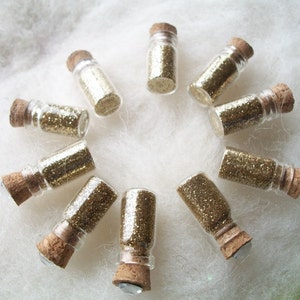 May include: Ten small glass bottles with cork tops filled with gold glitter. The bottles are arranged in a circle on a white fluffy surface.
