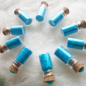 May include: Ten small glass bottles with cork stoppers filled with blue glitter. The bottles are arranged in a circle on a white fluffy surface.
