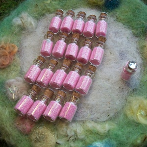 May include: A collection of small glass bottles filled with pink glitter. The bottles are arranged in a row on a fluffy white and green surface.