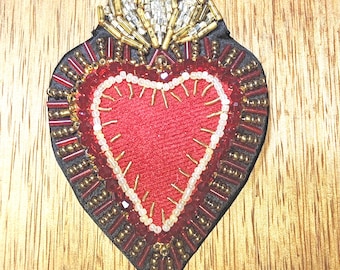 Pode incluir: Um patch bordado em forma de coração com um centro vermelho e um contorno branco. O patch é adornado com contas douradas e vermelhas e uma borda preta. A parte superior do coração apresenta um design em forma de coroa com detalhes prateados e dourados. Em fundo de madeira.