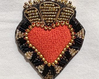 Könnte beinhalten: Herzförmige Brosche mit strukturiertem orangefarbenem Mittelteil, umrahmt von goldenen Perlen. Die Brosche hat schwarze und goldene Perlenakzente und ein goldenes Perlenkronen-Design oben. Wahrscheinlich handgefertigt.
