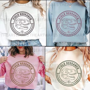 May include: Four sweatshirts in white, light blue, pink, and beige, each featuring a circular graphic with the text "FIELD RESEARCH" and a dinosaur skull. The design is in black, burgundy, brown, and green.