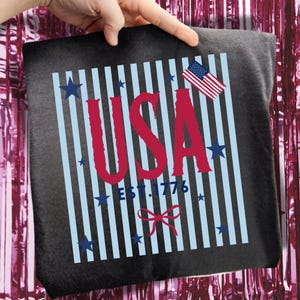 May include: A dark gray t-shirt featuring a patriotic design. The design includes the word "USA" in red, with light blue vertical stripes, blue stars, and an American flag. The text "EST. 1776" is also present.