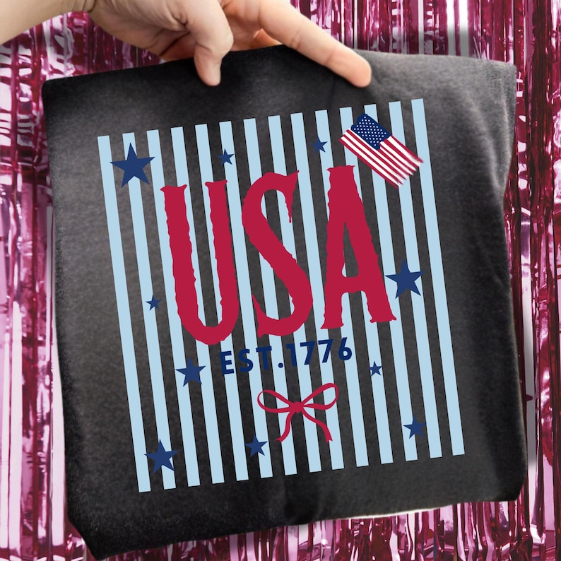 May include: A dark gray t-shirt featuring a patriotic design. The design includes the word "USA" in red, with light blue vertical stripes, blue stars, and an American flag. The text "EST. 1776" is also present.
