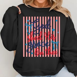 May include: Black sweatshirt with a vertical striped design in pink and white. The text "God Shed His Grace On Thee" is in blue and red script, with blue bows and stars.
