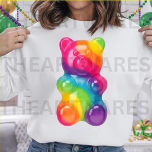 May include: White sweatshirt featuring a large, colorful gummy bear design. The gummy bear is a vibrant mix of rainbow colors, including pink, orange, yellow, green, blue, and purple. The text "HEART WARES" is printed on the sweatshirt.