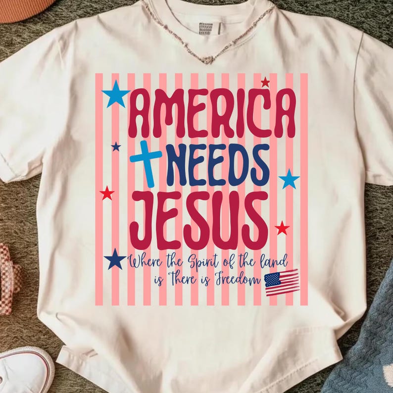 May include: Cream-colored t-shirt with the text "AMERICA NEEDS JESUS" in red and blue, accented with stars and stripes. The shirt also features the phrase "Where the Spirit of the land is There is Freedom" and a small American flag.