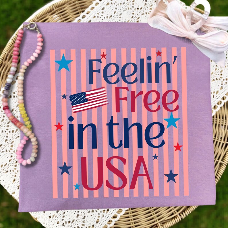 May include: A lilac-colored t-shirt with the phrase "Feelin' Free in the USA" in navy blue and red lettering, along with an American flag graphic. The design includes pink vertical stripes and blue and red stars.