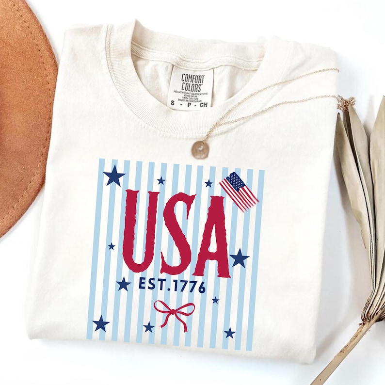 May include: Cream-colored t-shirt with a patriotic USA design. The design features the word "USA" in red, blue stars, blue stripes, a small American flag, and the text "EST. 1776". A gold necklace is draped across the shirt.