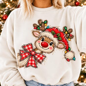 May include: A cream-colored crewneck sweatshirt featuring a graphic of a reindeer with a red nose, wearing a red Santa hat with holly and colorful Christmas lights on its antlers. A large red and white checkered bow is tied at the neck.
