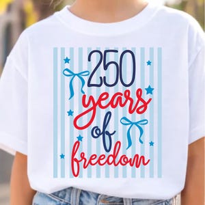May include: White t-shirt with a patriotic design. The design features the number "250" in navy blue, the words "years of freedom" in red script, and blue bows and stars. The background has light blue vertical stripes.