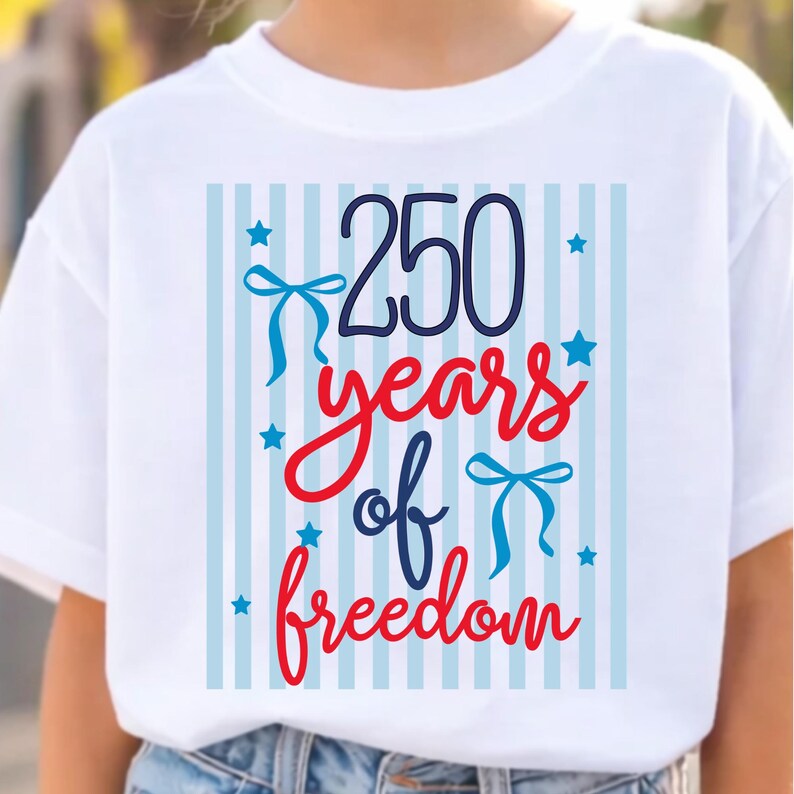 May include: White t-shirt with a patriotic design. The design features the number "250" in navy blue, the words "years of freedom" in red script, and blue bows and stars. The background has light blue vertical stripes.