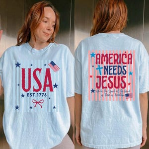 May include: Light blue t-shirts with patriotic designs. One shirt features "USA EST. 1776" with stars, a bow, and an American flag. The other shirt says "AMERICA NEEDS JESUS" with stars and an American flag.