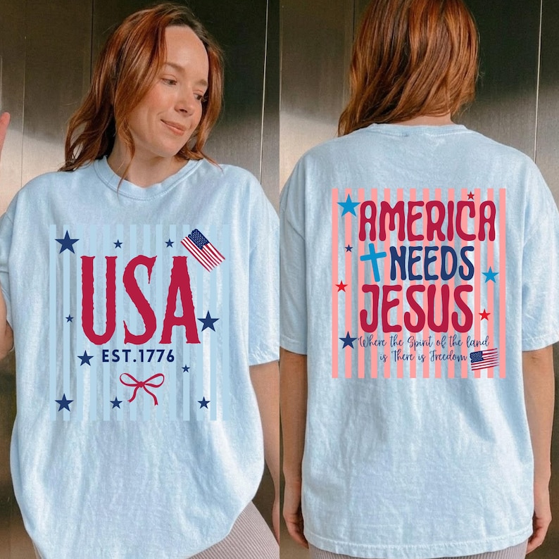 May include: Light blue t-shirts with patriotic designs. One shirt features "USA EST. 1776" with stars, a bow, and an American flag. The other shirt says "AMERICA NEEDS JESUS" with stars and an American flag.
