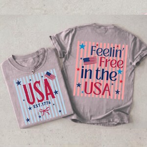 May include: Two gray t-shirts with patriotic designs. One shirt features the word "USA" in red with blue stripes and stars, and the text "EST.1776". The other shirt says "Feelin' Free in the USA" with a US flag.