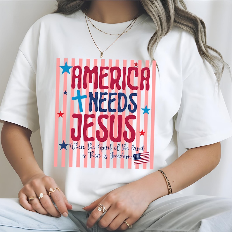 May include: White t-shirt featuring the text "AMERICA NEEDS JESUS" in red and blue, with a cross and stars. The design includes the phrase "Where the Spirit of the land is There is Freedom" and a small American flag.
