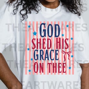 May include: White t-shirt with a patriotic design. The design features the words "GOD SHED HIS GRACE ON THEE" in blue and red, with blue stars and a red bow. The background has red and white stripes.