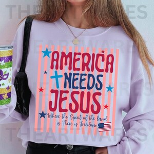May include: A light purple sweatshirt with the text "AMERICA NEEDS JESUS" in red and blue, with a cross and stars. Below the text is the phrase "Where the Spirit of the land is There is Freedom" and an American flag.