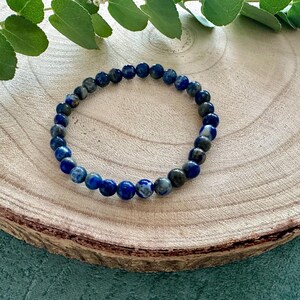 K&ouml;nnte beinhalten: Ein blaues und graues Edelstein-Armband mit runden Perlen. Das Armband liegt auf einer Holzoberfl&auml;che.