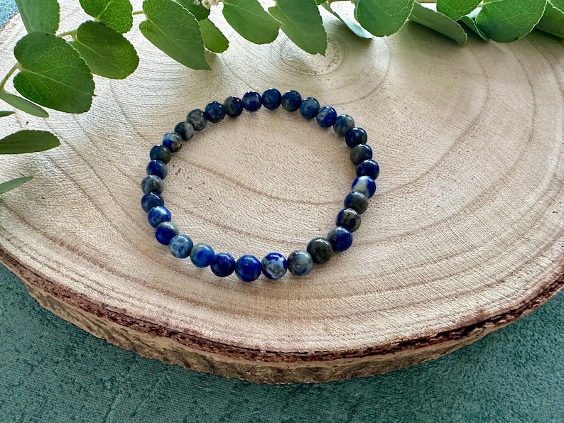 K&ouml;nnte beinhalten: Ein blaues und graues Edelstein-Armband mit runden Perlen. Das Armband liegt auf einer Holzoberfl&auml;che.