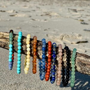 K&ouml;nnte beinhalten: Eine Sammlung von 10 bunten Perlenarmb&auml;ndern, jedes mit einer anderen Farbe und Textur, auf einem St&uuml;ck Treibholz ausgestellt. Die Armb&auml;nder sind aus nat&uuml;rlichen Steinen gefertigt, darunter T&uuml;rkis, Lapislazuli und Jade.