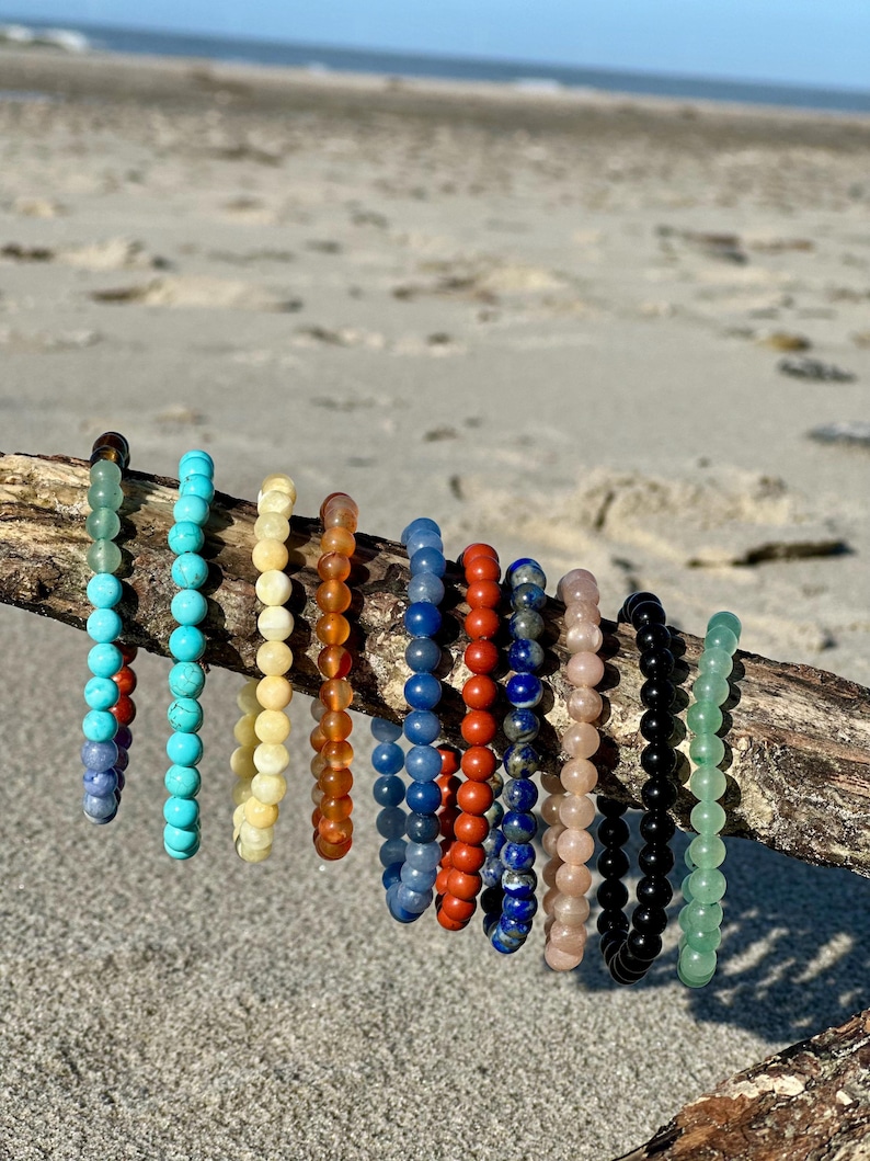 K&ouml;nnte beinhalten: Eine Sammlung von 10 bunten Perlenarmb&auml;ndern, jedes mit einer anderen Farbe und Textur, auf einem St&uuml;ck Treibholz ausgestellt. Die Armb&auml;nder sind aus nat&uuml;rlichen Steinen gefertigt, darunter T&uuml;rkis, Lapislazuli und Jade.