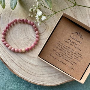 Könnte beinhalten: Ein rosafarbenes Rhodonit-Armband mit einer braunen Schachtel, auf der "SjælRokk Rhodanit Stein der Heilung" steht und die heilenden Eigenschaften des Steins beschreibt.