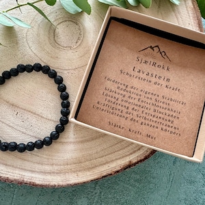 Könnte beinhalten: Ein schwarzes Lava-Stein-Armband in einer kleinen Box mit einer Beschreibungskarte. Die Karte lautet "SjælRokk Lavastein Schutzstein der Kraft" und listet die Vorteile des Tragens von Lavastein auf, darunter Erdung, Stabilität und Stressabbau.