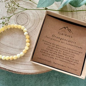 Könnte beinhalten: Ein gelborangenes Calcit-Armband mit einer kleinen Schachtel, die Informationen über die Eigenschaften des Steins enthält. Die Schachtel trägt die Aufschrift "SjælRokk Energiestein der Lebensfreude Orangencalcit" und listet die Vorteile des Steins auf, darunter die Förderung von positiver Energie, Kreativität und Selbstheilung.